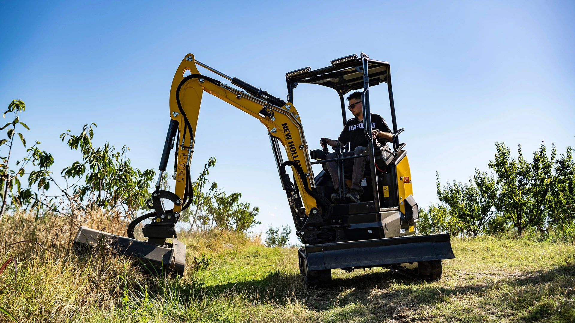Operator in New Holland mini crawler excavator, working in field with clear blue sky backdrop.