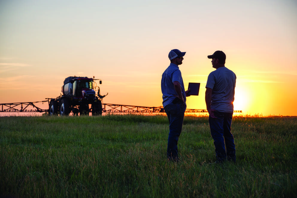 Two farmers discuss a tablet in a field at sunset, with a crop sprayer tractor behind them and golden light creating a hopeful mood.