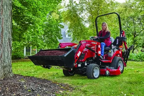 A person operates a Case IH subcompact tractor, loaded with mulch, on a grassy lawn surrounded by trees, near a house with a visible window.