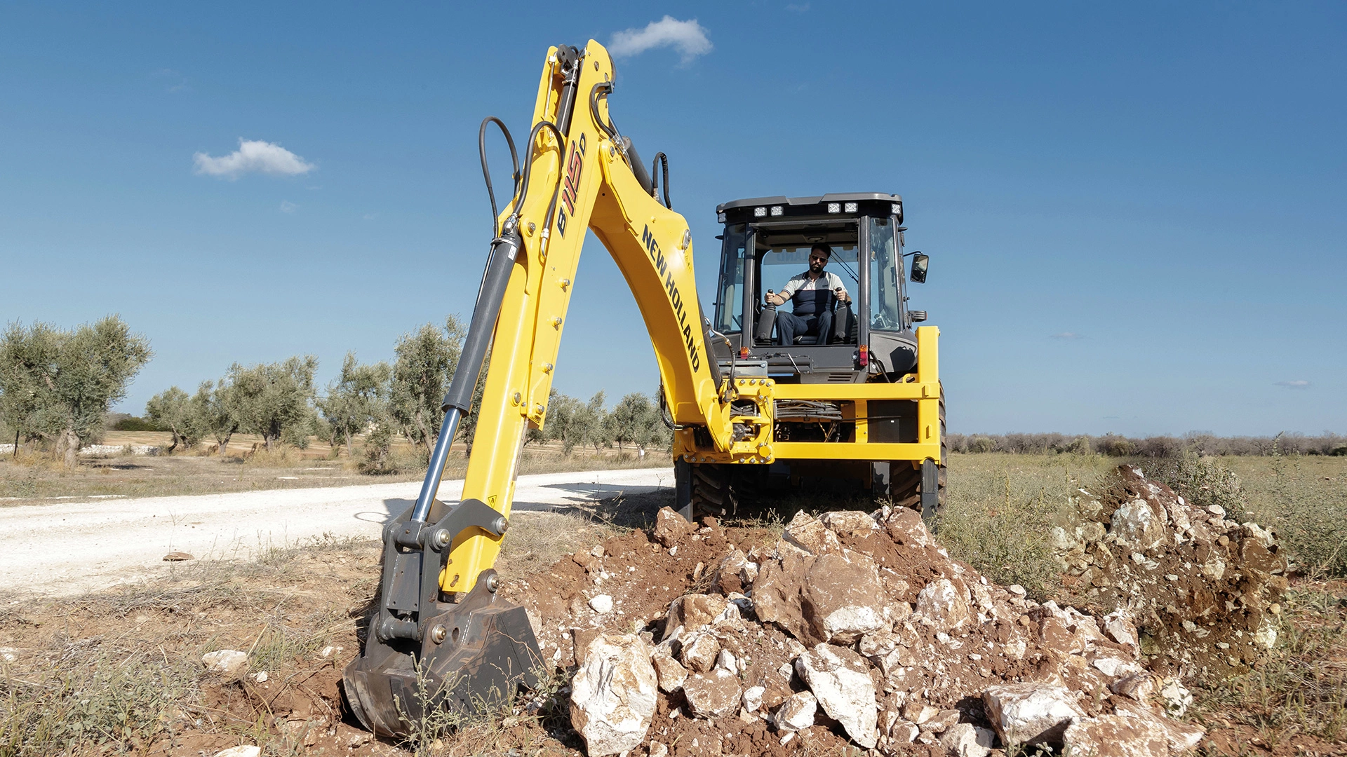 New Holland Backhoe at work, digging in rocky terrain, clear sky, operator in cabin, olive trees in background.
