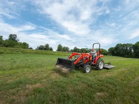 A person drives a Case IH Farmall Compact 35A tractor across a green field under a bright blue sky with scattered clouds. The scene conveys a sense of calm and productivity.