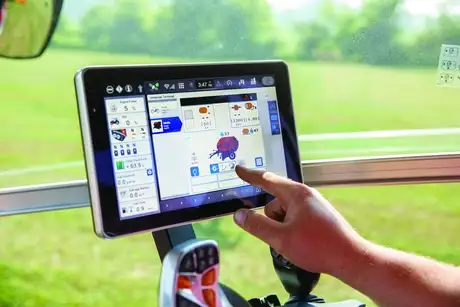 A close-up of a hand interacting with a FieldOps touchscreen inside a vehicle, displaying agricultural control panels. A green field is visible through the window.