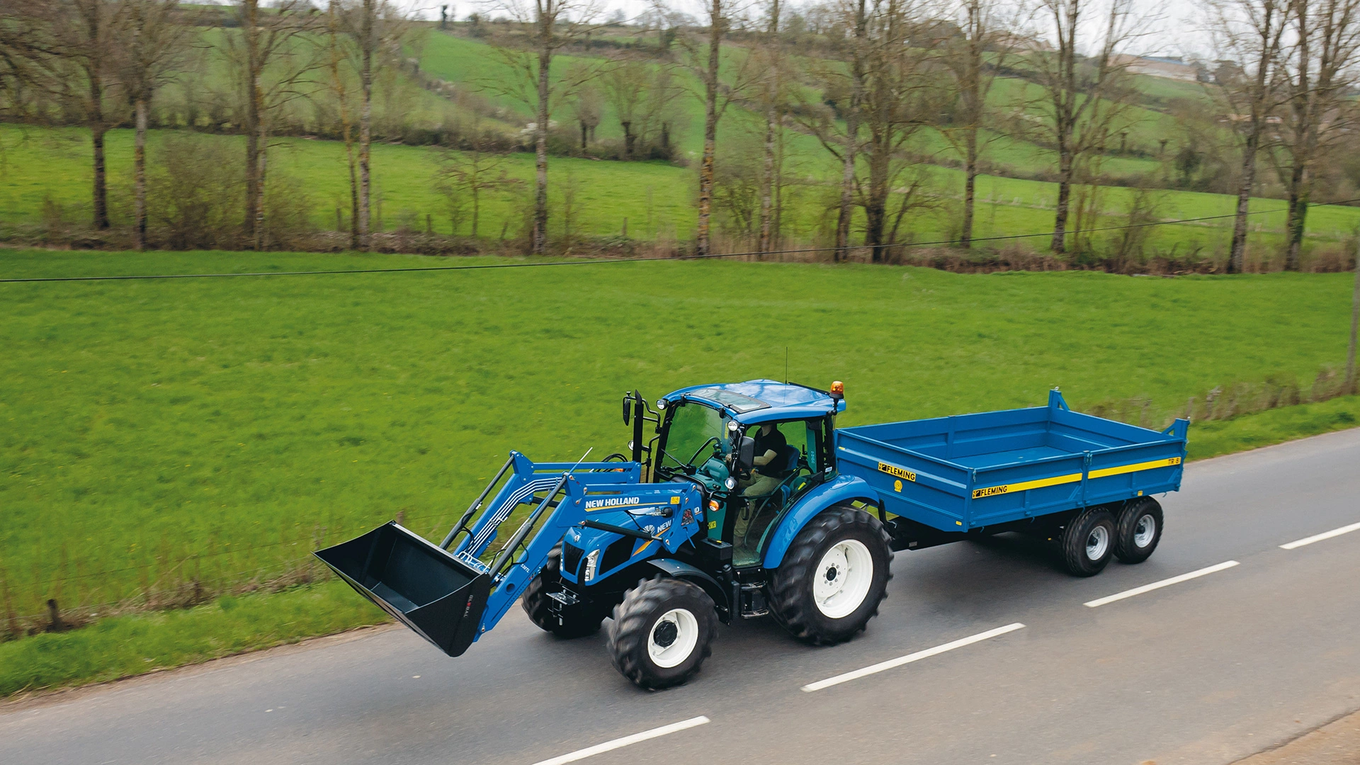 A blue New Holland tractor with a front loader attachment driving on a road, pulling a blue Fleming trailer, with a lush green field and trees in the background.