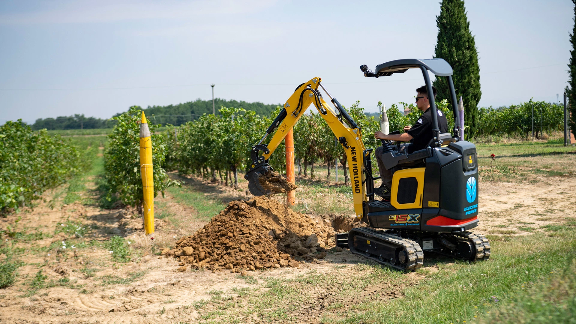 Operator in New Holland mini crawler excavator, working in field with clear blue sky.