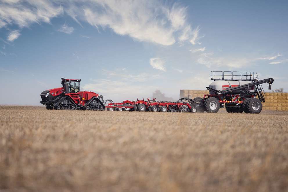 Red Case IH tractor with tracks pulls large farming equipment across dry field under blue sky with wispy clouds.