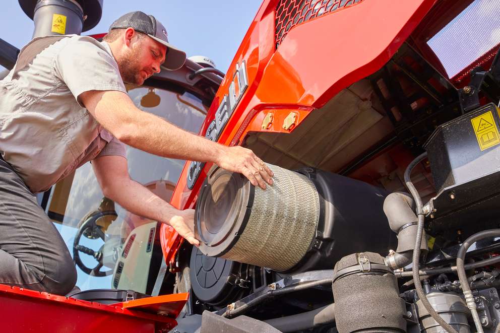 A man in a cap replaces or inspects the cylindrical air filter of a red Case IH tractor engine.