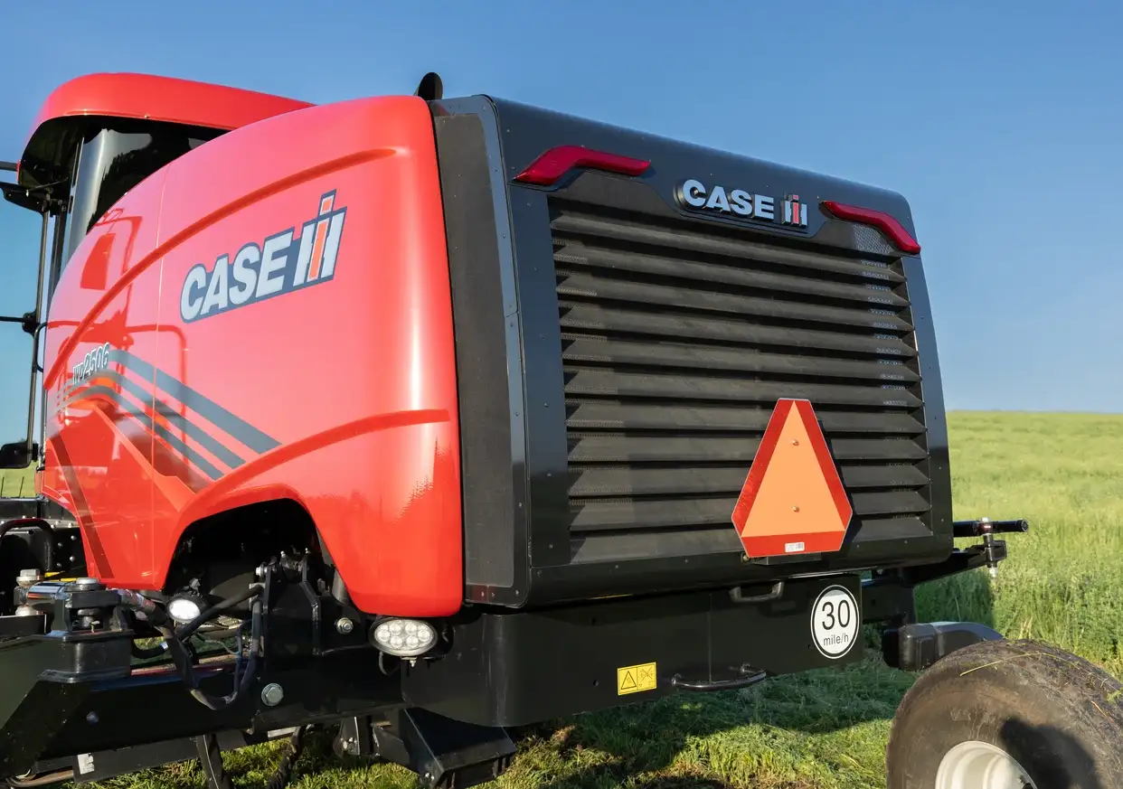 Close-up of a red CASE IH agricultural baler in a sunny green field, showing its black grille, slow-moving vehicle sign, and large tire.