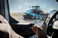 A person inside a vehicle holding a smartphone displaying a tractor monitoring app, with a blurred tractor visible through the window and digital connectivity icons floating above it.