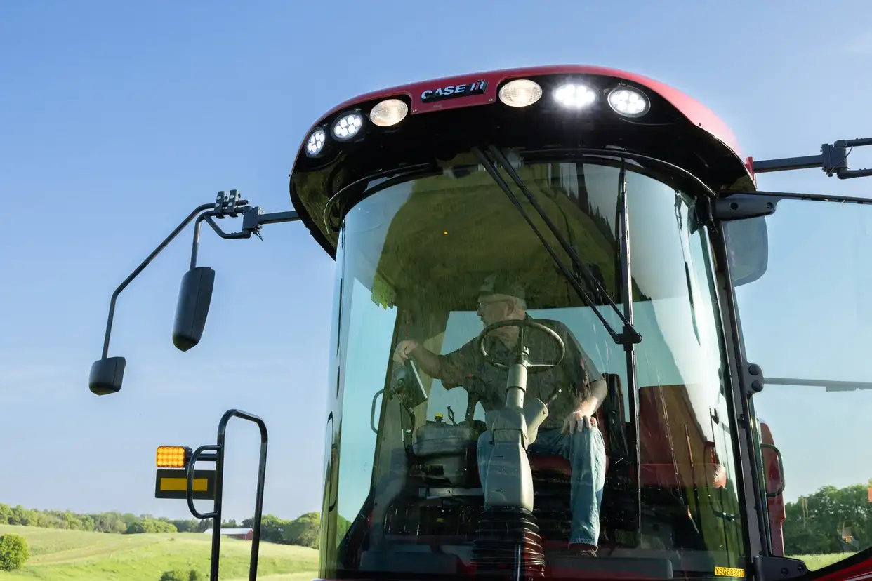 Front view of a red Case IH combine harvester with curved windshield and work lights, a farmer inside, and green fields beneath a clear sky.