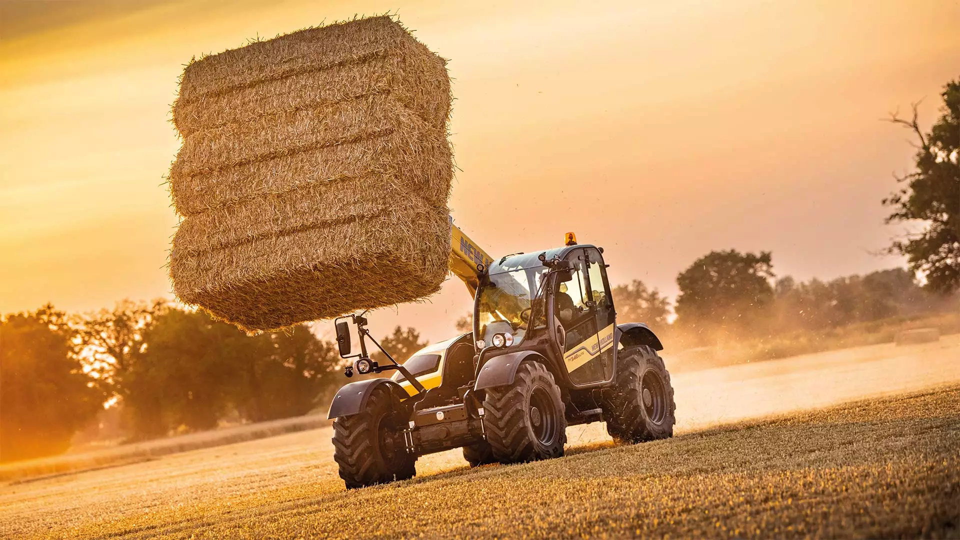 Agricultural telehandler lifting large hay bale at sunset in a farm field.