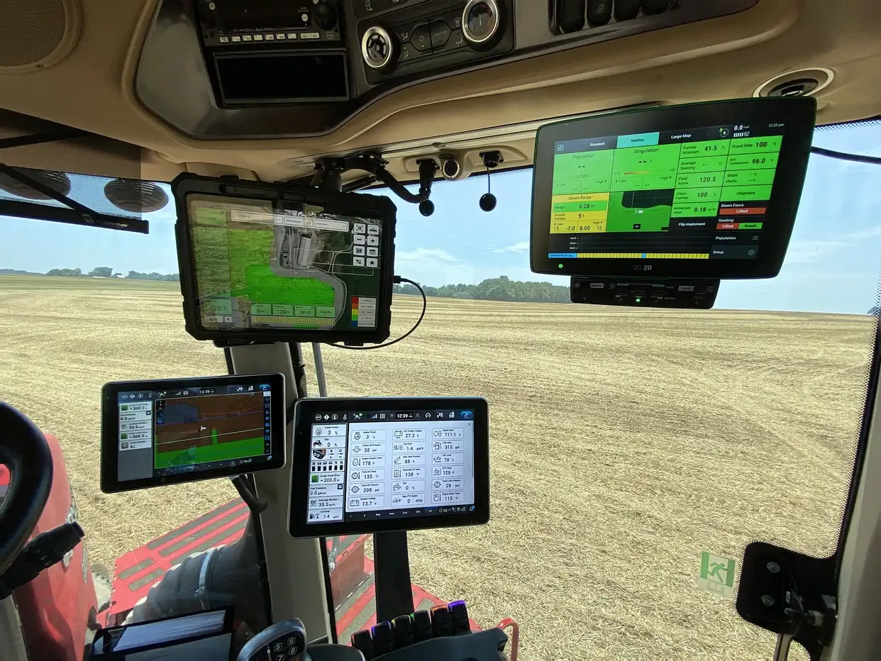Interior of a tractor cab with multiple digital screens displaying farming and GPS data. Expansive field visible through the window, suggesting a high-tech agriculture setting.