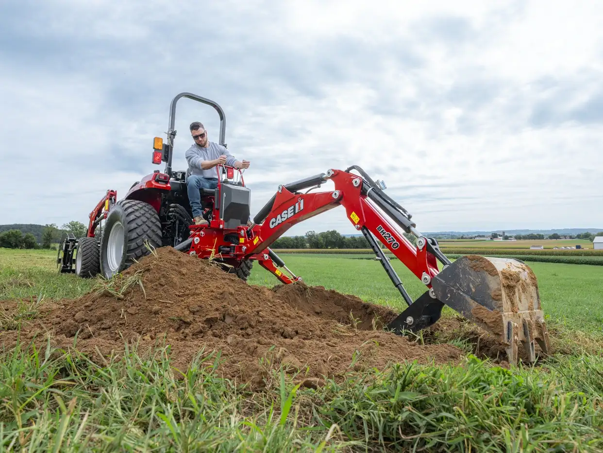 Farmer operates a red Case IH tractor with backhoe, digging a mound of soil in a green field beneath a cloudy sky.