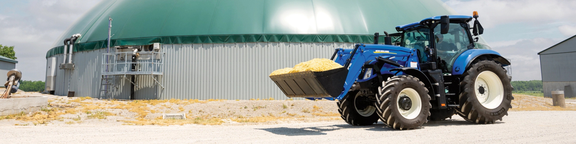 New Holland tractor with a loader carries bucket of material across the farm.
