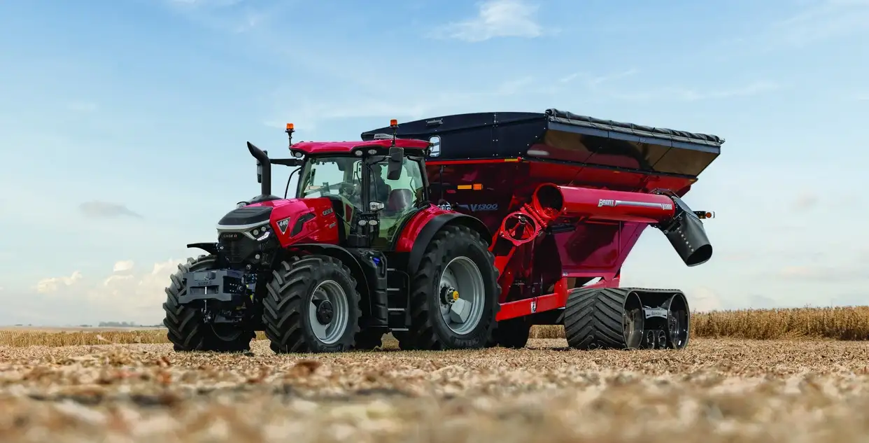 A Case IH Optum tractor pulls a matching grain cart across a vast, flat field under a bright blue sky with scattered clouds. The scene appears calm and industrious.