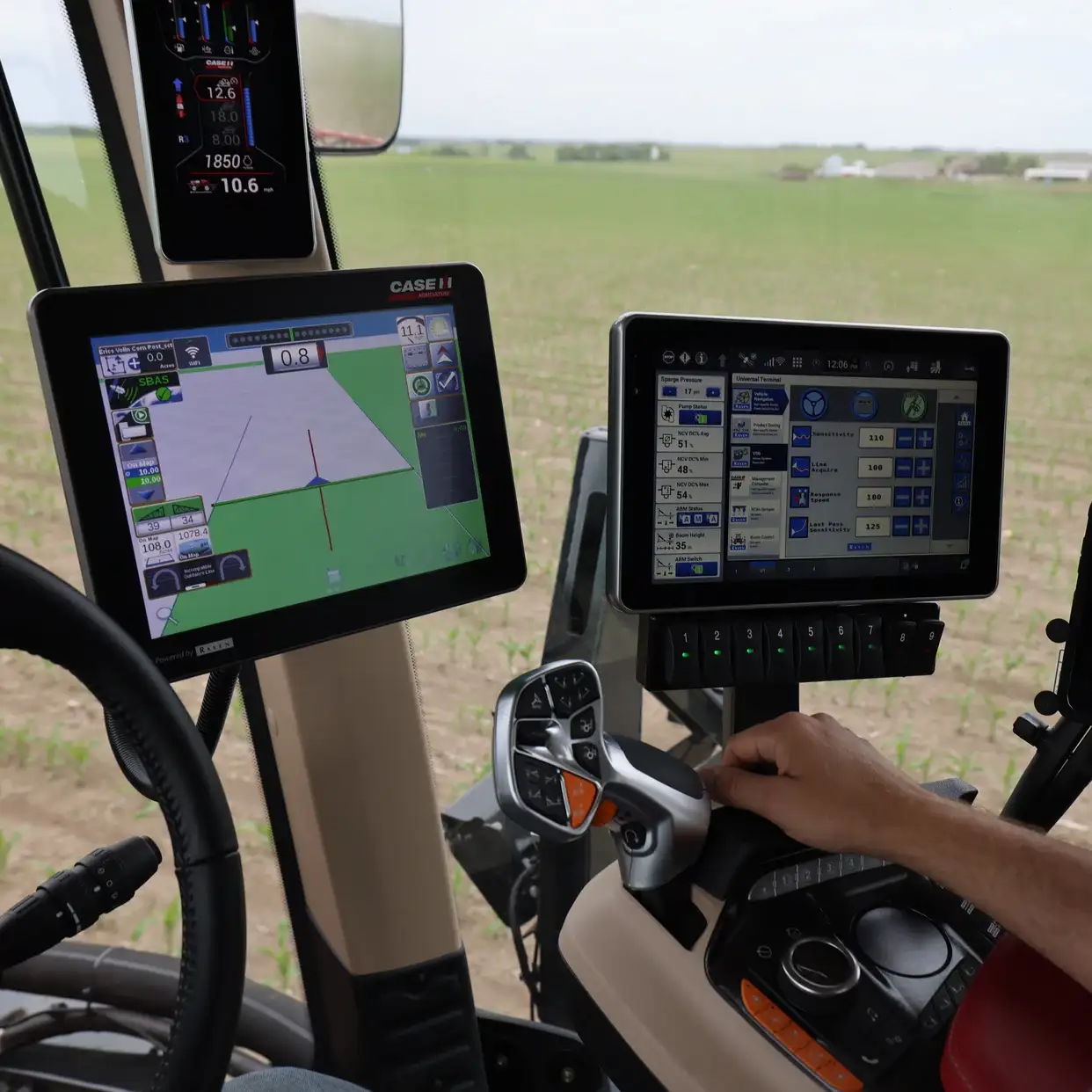 View from inside a tractor cabin showing two touchscreens displaying Slingshot, steering wheel, and a hand operating controls. Background of a vast green field.