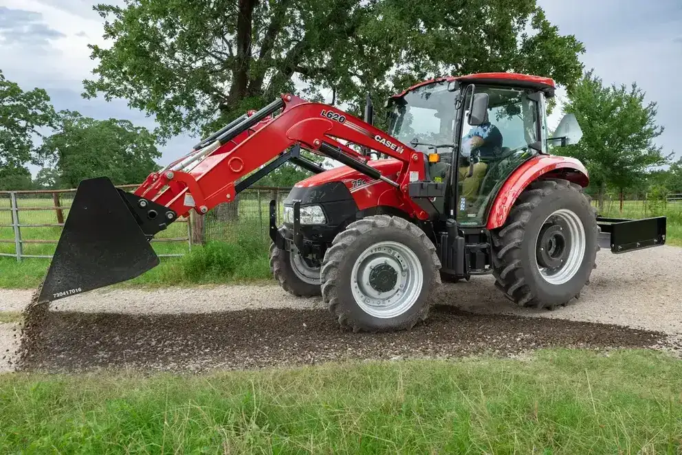 A Case IH tractor with a front loader scatters gravel on a dirt path in a green field.