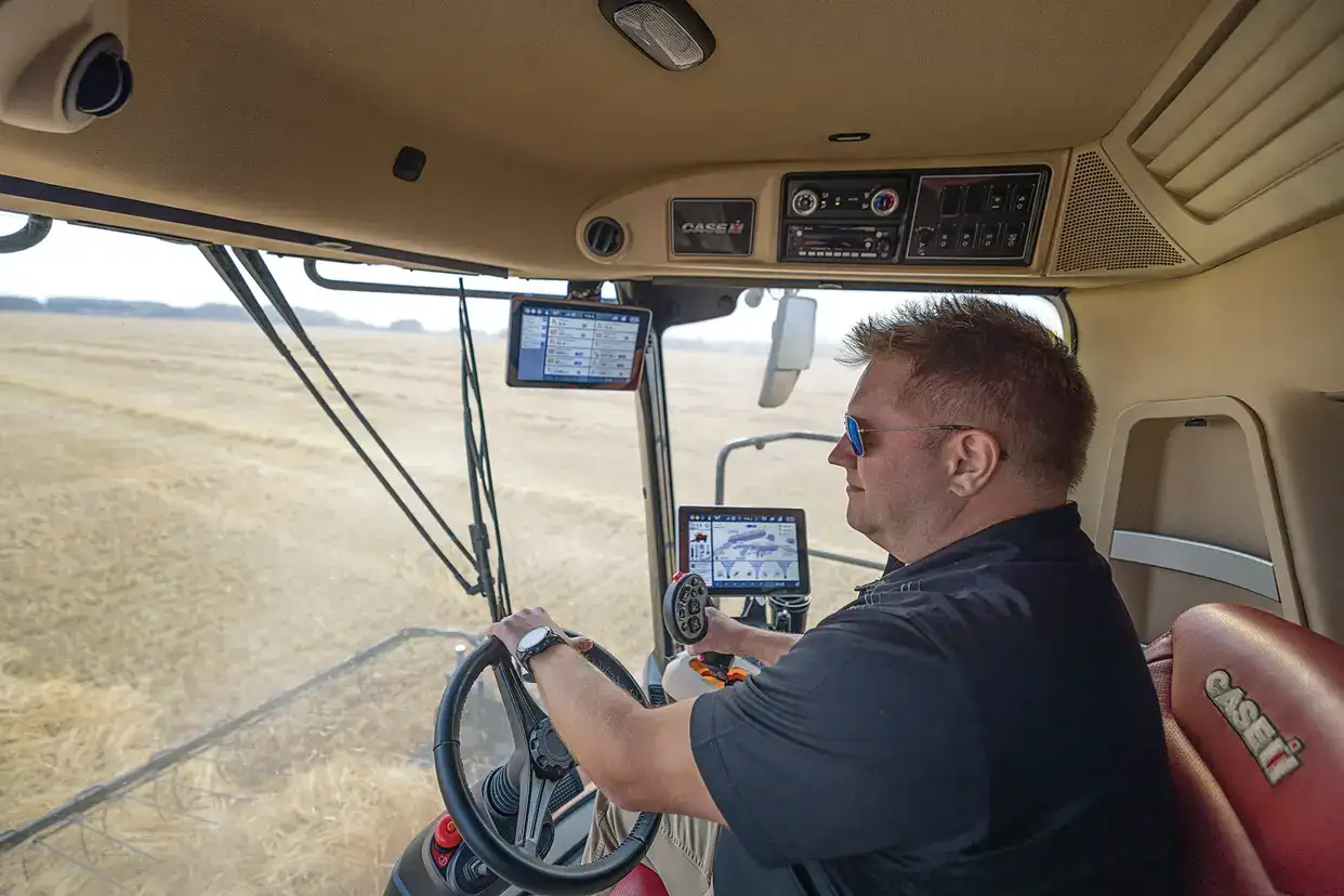 A man drives a Case IH combine harvester across a dry field, focused on digital monitors and controls inside the cab on a sunny day.