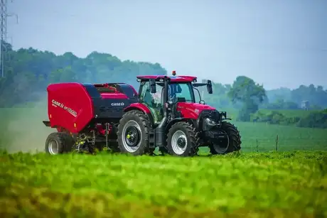A Case IH tractor with a hay baler attachment operates in a lush, green field under a clear sky. 