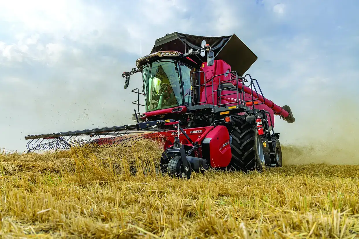 A red Case IH combine harvester works in a golden wheat field under a partly cloudy sky. Dust trails behind, conveying a sense of motion and productivity.