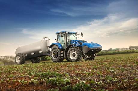 foto ao ar livre, do trator T6 Methane Power da New Holland trabalhando em um vasto campo. O trator está puxando um pulverizador agrícola como implemento.