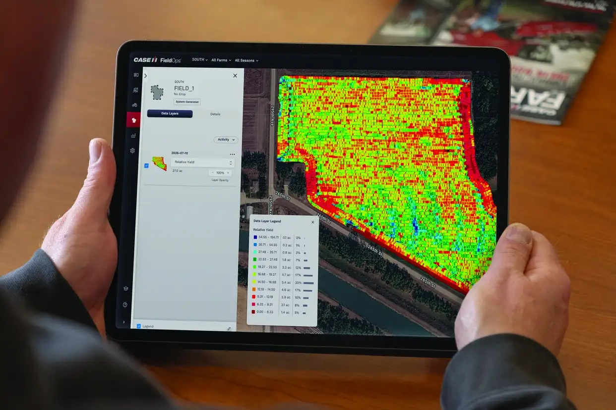Hands hold a tablet showing a precision agriculture field map with red, yellow, and green data zones, legend, and satellite imagery.