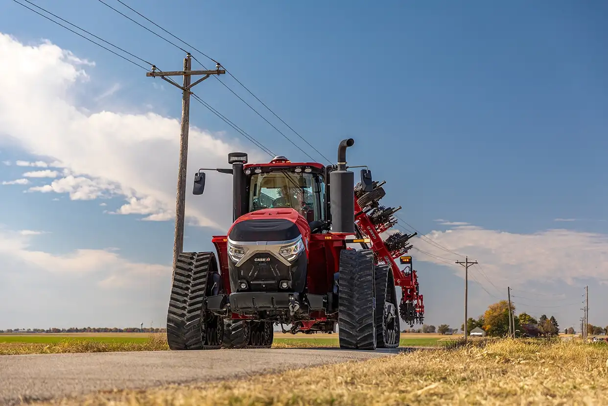 A red Case IH tractor with tracks drives on a rural road, with a narrow and folded Nutri-Tiller 1000 series strip tillage tool attached. 
