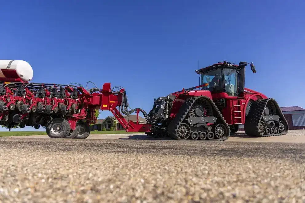 Red Case IH tractor with large treads towing agricultural equipment on a gravel pathway. Clear blue sky and a barn in the background convey a bright, rural scene.