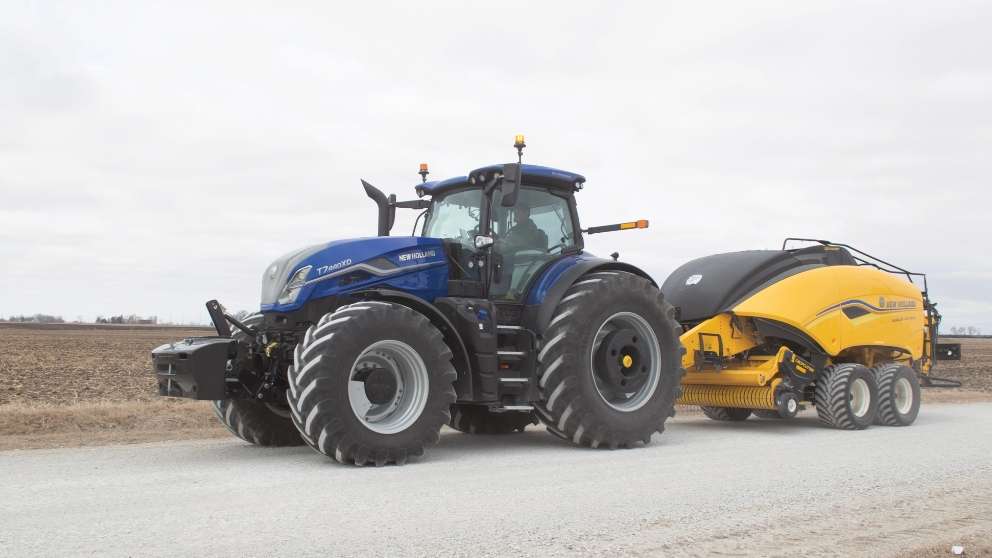 Blue tractor pulls yellow hay baler on a gravel road.