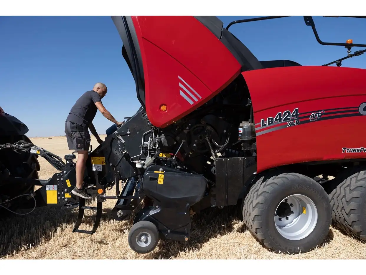 Technician performing maintenance on a Case IH LB424 XLD AFS large square baler with the hood open in a stubble field