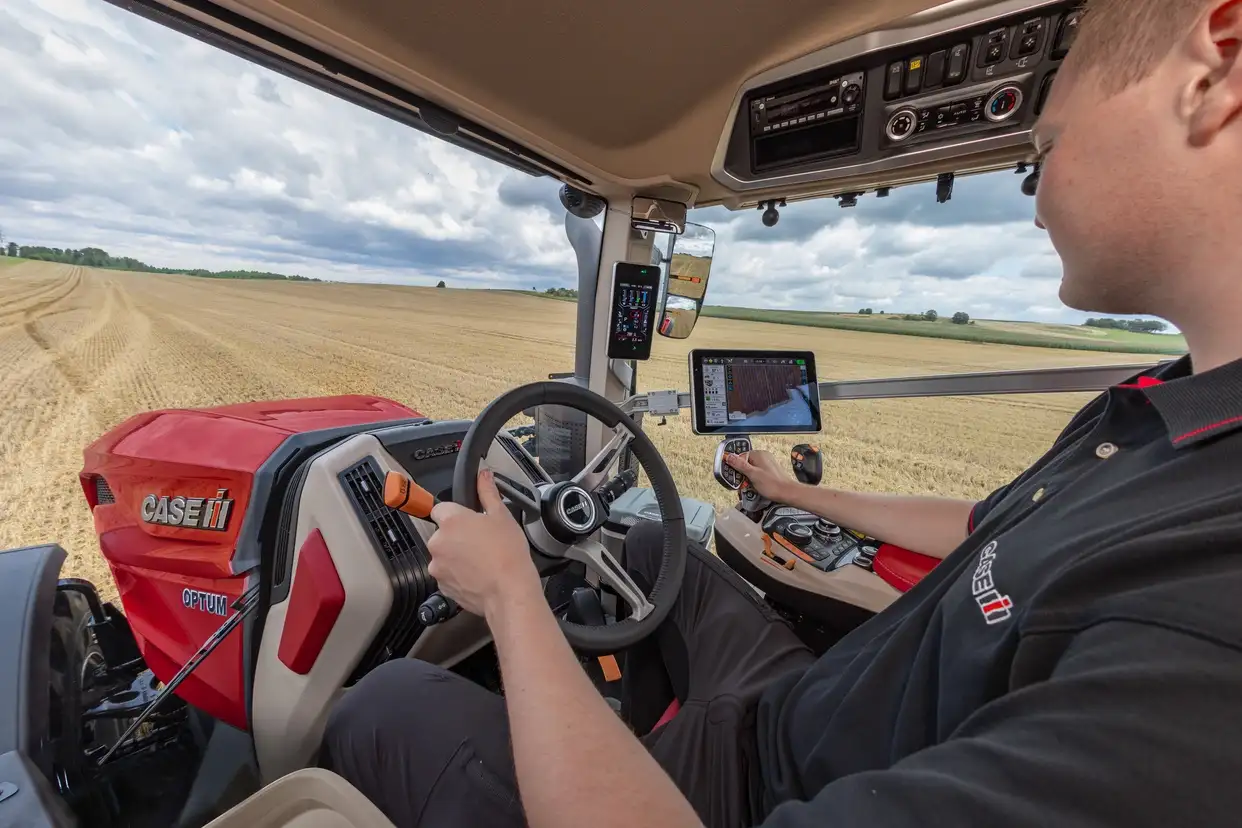 A farmer steers a red Case IH tractor through a harvested field, checking the in-cab GPS display under a cloudy sky.