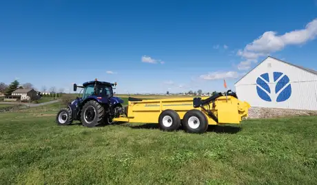 A tractor pulls a yellow box spreader in front of a barn.