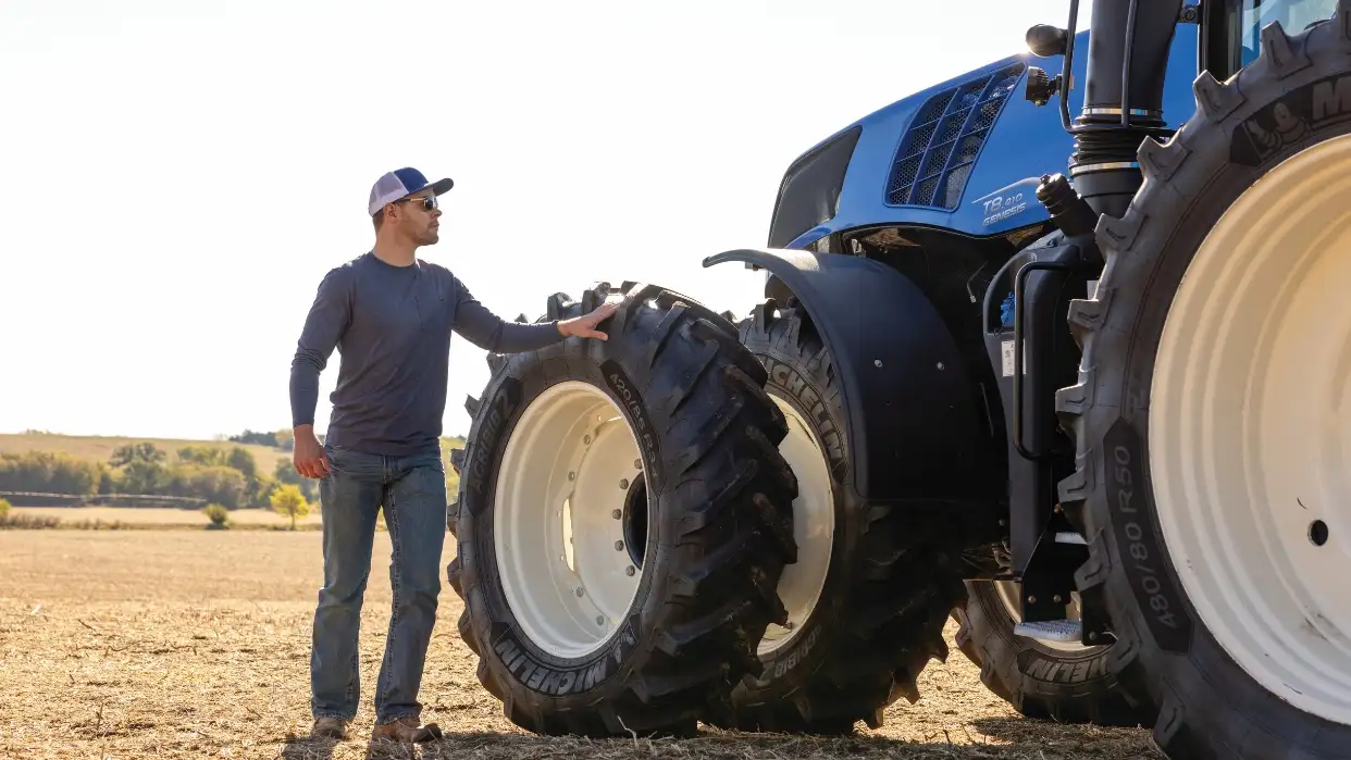 Man in a cap stands next to a big blue tractor.