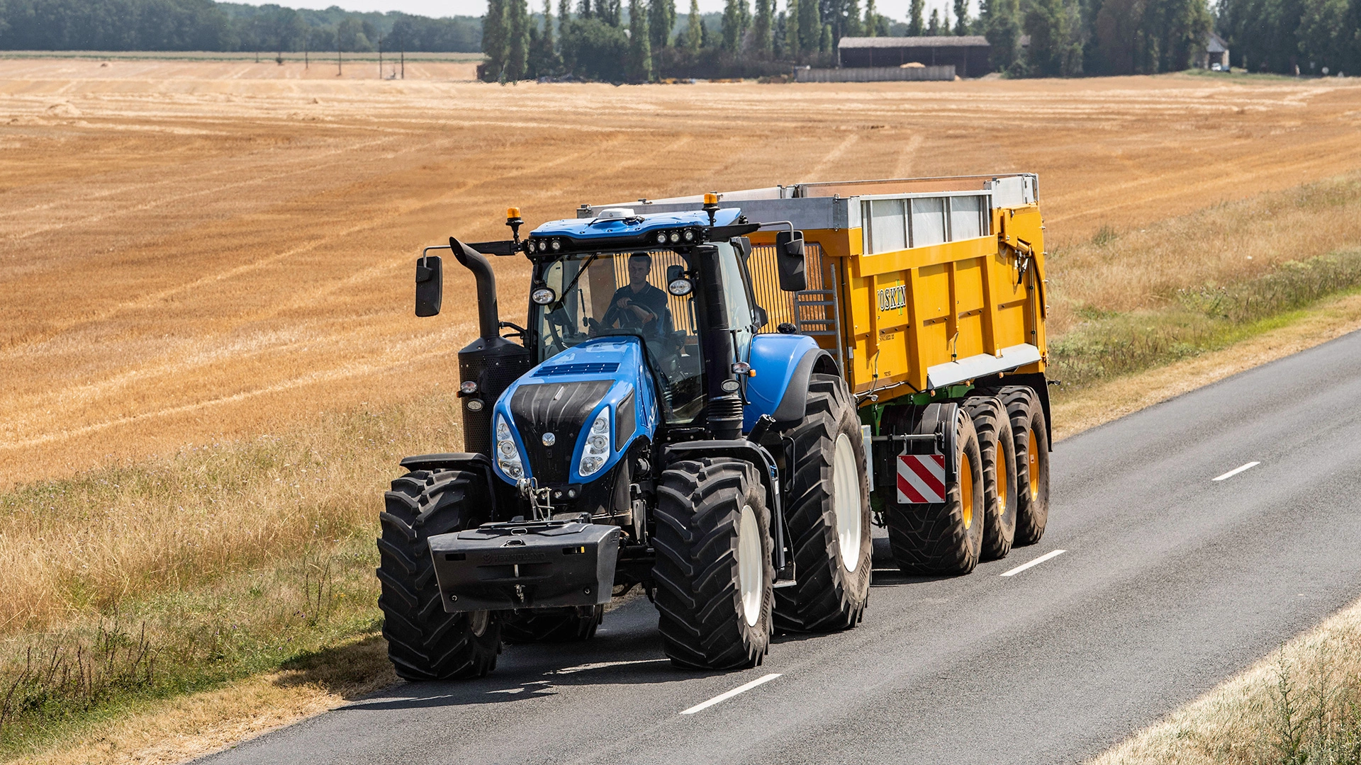 New Holland Tractor on road with trailer, golden field backdrop, driver visible in cab.
