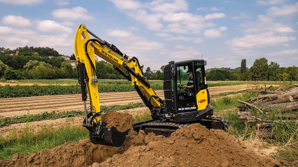New Holland mini crawler excavator digging soil in agricultural field, village and trees in background.