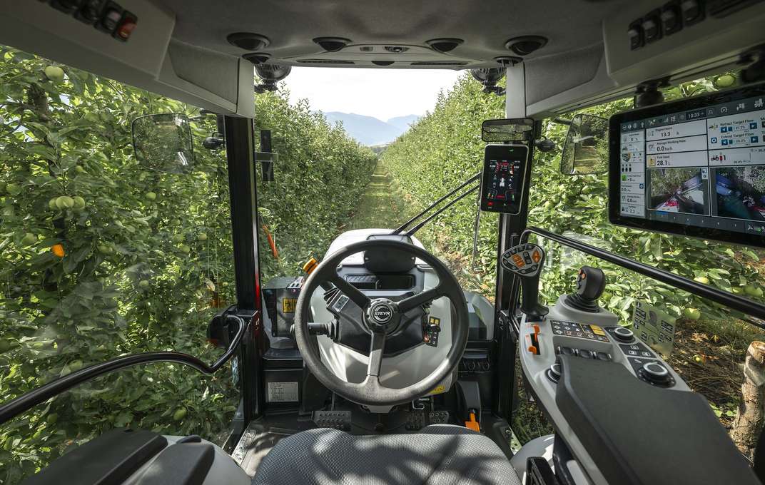STEYR Spezial 4120F orchard tractor towing an orange sprayer along a gravel track between apple orchard rows, mountains behind
