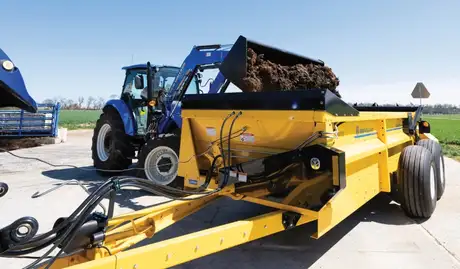 A blue tractor dumps manure into a yellow box spreader.