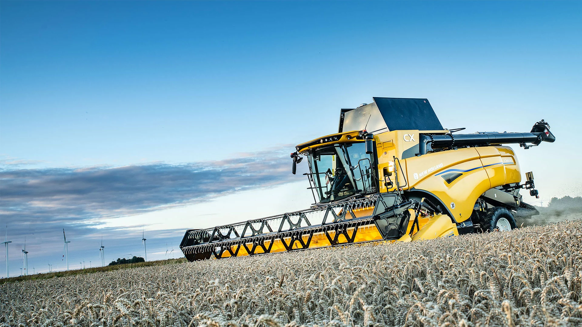 New Holland CX7 & CX8 Combine Harvester in field, under a blue sky with wind turbines in background.