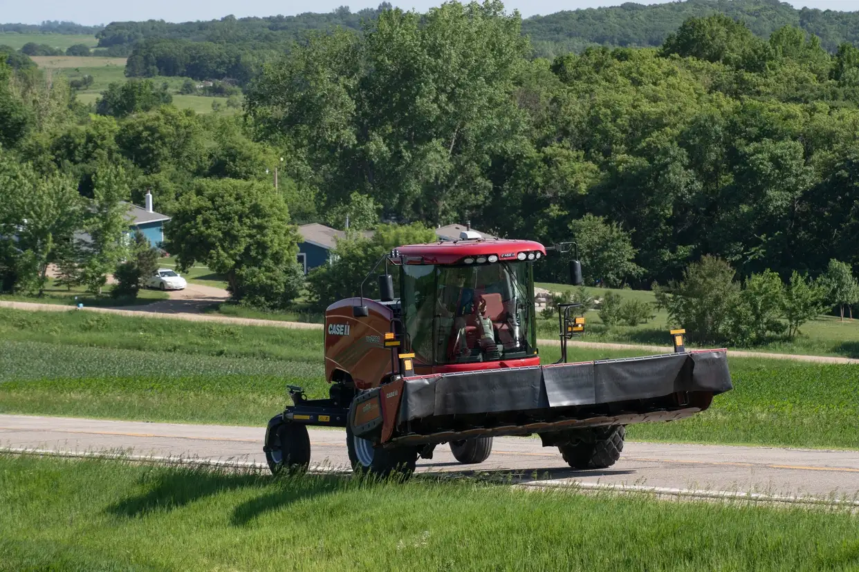 Case IH hay mower with an operator cuts a grassy field beside a rural road, surrounded by trees, farmland, and rolling hills.