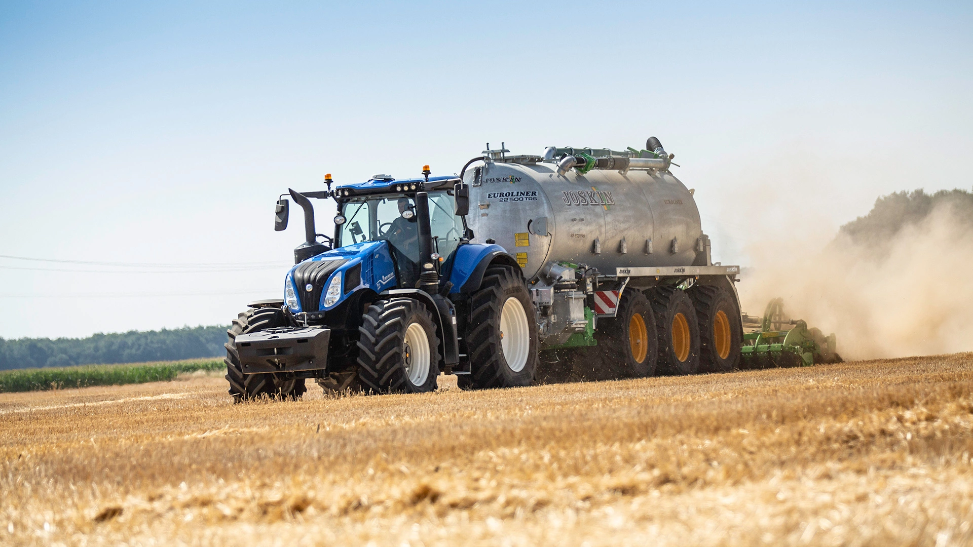 New Holland Tractor T8 Genesis with trailer in a field, harvesting crops, creating dust trail.