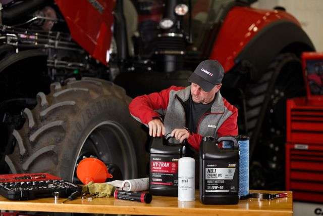 A mechanic in a Case IH branded jacket and cap opens a bottle of Hy-Tran oil on a workbench with engine oil, filters, and tools near a red tractor.