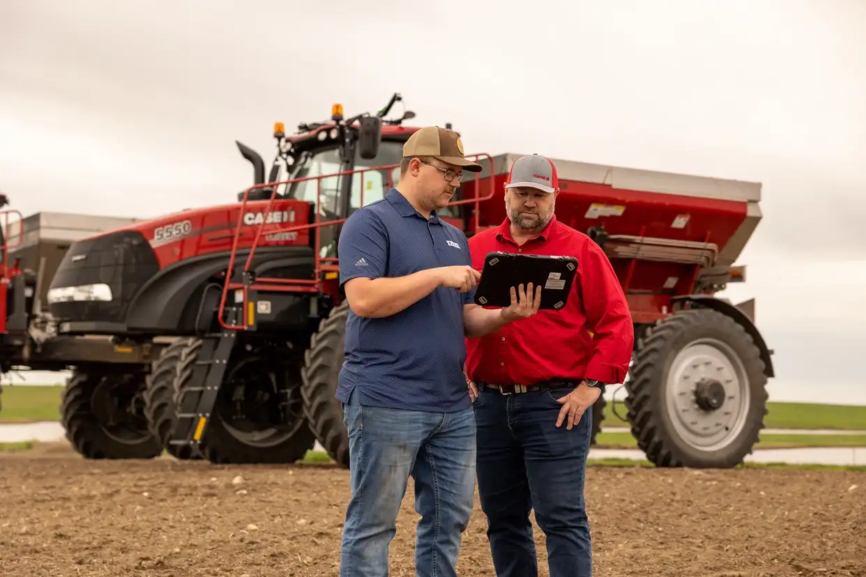 Two farmers stand in a field beside red farming machinery, discussing data on a tablet. The scene is overcast, conveying a focused, collaborative atmosphere.