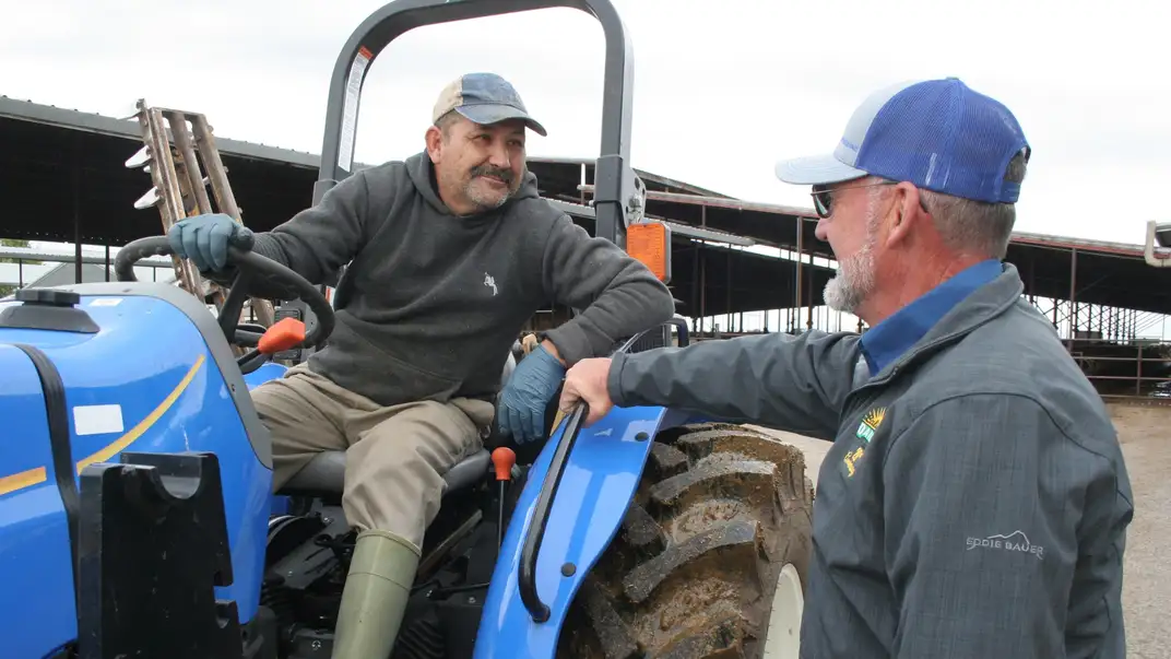 two men smiling at each other, one seated in a tractor, the other standing beside him.