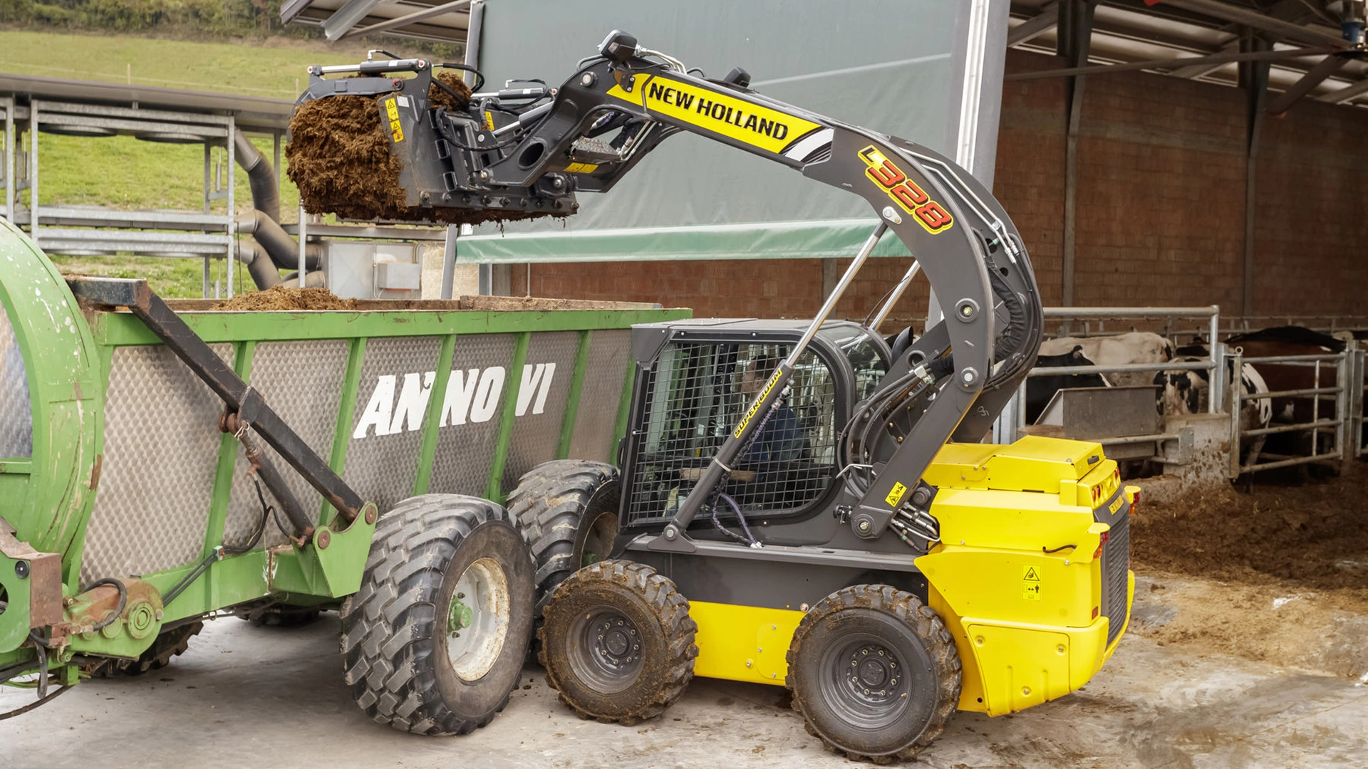 New Holland skid steer loader operating at a farm, lifting a load of material into a green trailer.