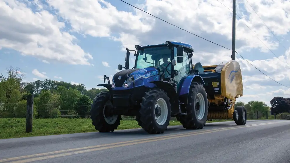 Blue tractor pulling yellow hay baler on a road.