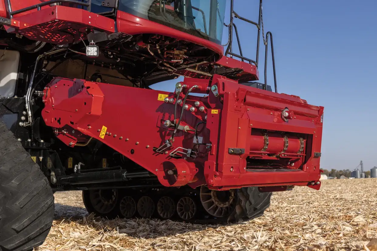 A close-up view of a red combine harvester feeder house, showing hydraulic lines, safety decals, and heavy-duty tracks on a harvested corn field.