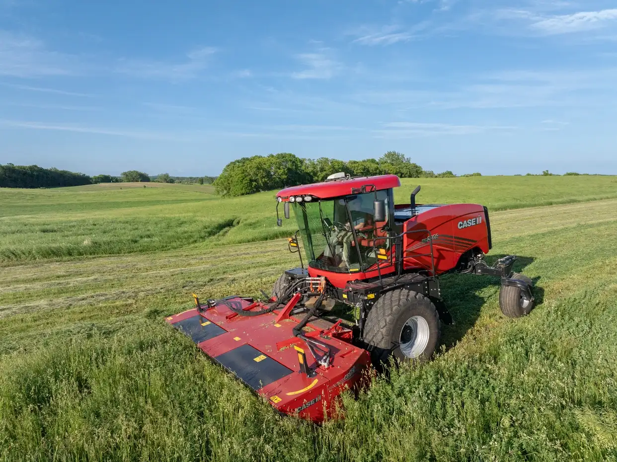 Red Case IH mower-conditioner cuts a green field beneath blue skies, with rolling farmland and trees creating a peaceful rural scene.