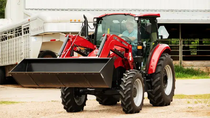 A Case IH tractor with large front loader parked in front of a metal barn. A person sits inside the cab. 