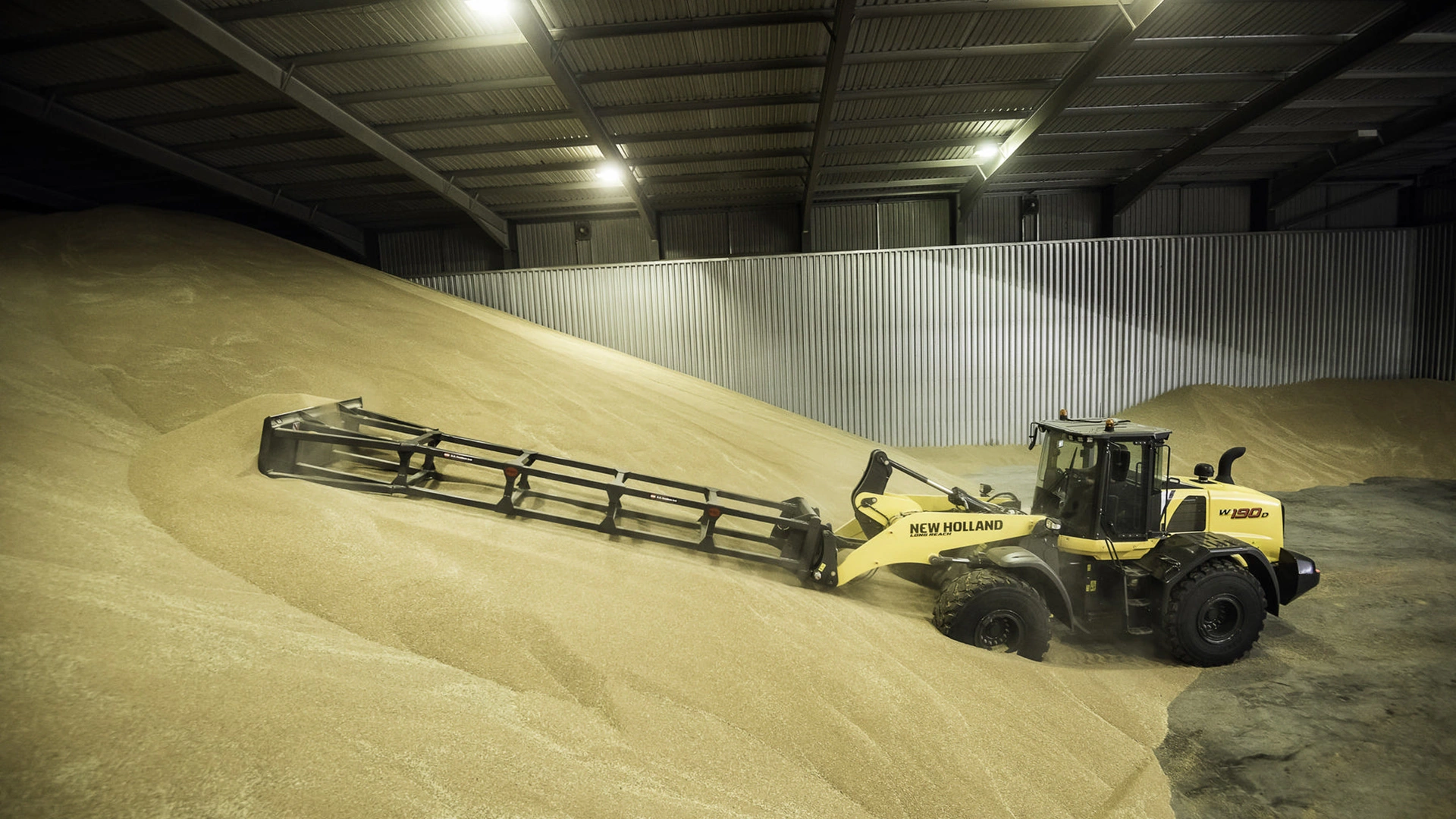 New Holland W190D wheel loader at work efficiently moving a large quantity of grain within an agricultural storage facility.