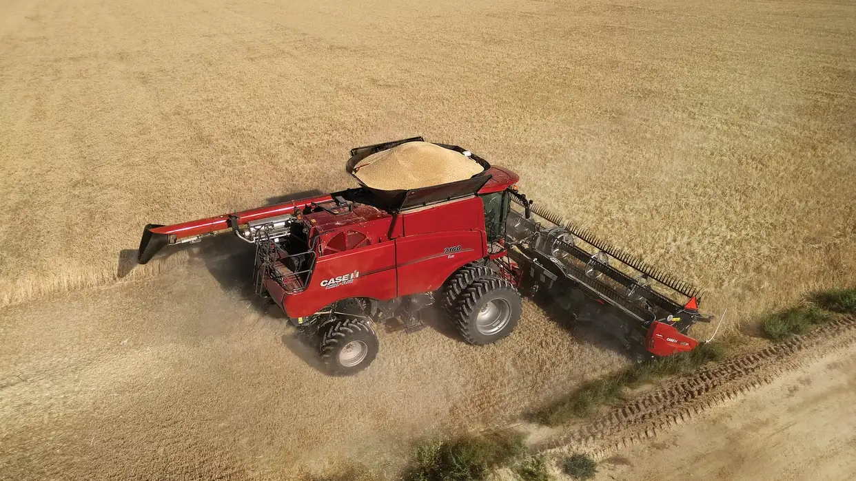 A red Case IH combine harvester cuts through a golden wheat field from above, raising a dusty cloud and leaving tire tracks behind.