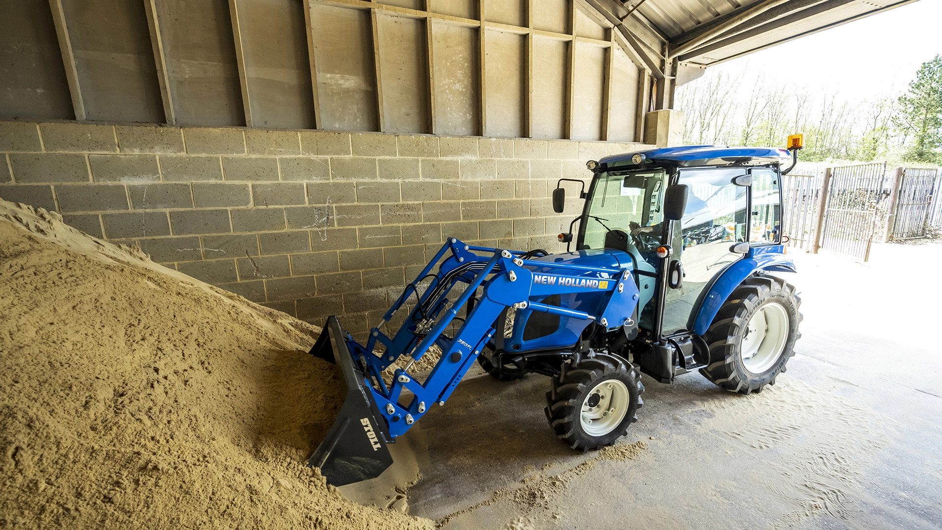 New Holland tractor with front loader bucket moving sand in an industrial storage area.